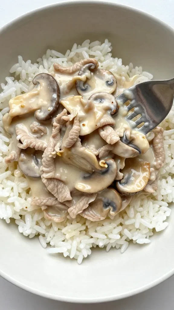 Tight overhead crop focusing on the textures: fork pulling through creamy mushroom pork on top of plain white rice, thin strips of pork, browned mushroom slices, smooth pale sauce with a slight Dijon tint, tiny garlic bits, light seasoning specks only, matte ceramic bowl, bright but diffused daylight, no additional ingredients or decorations