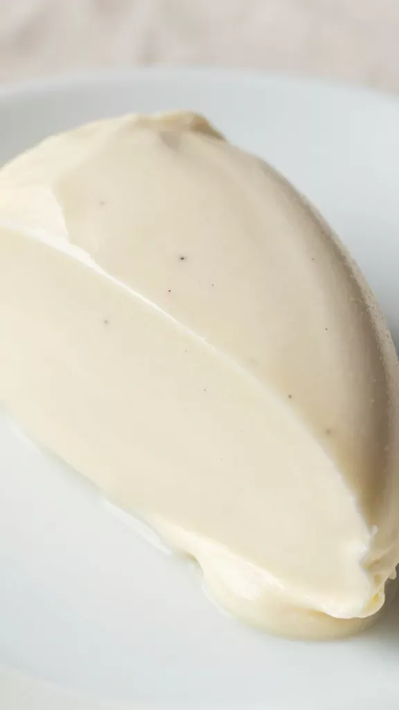 Extreme close-up of a thick slice of vanilla bean semifreddo on a simple white plate, creamy off-white surface with visible tiny black vanilla specks throughout, soft satin-like texture with slightly melty edges, clean slice lines, no toppings or sauces, natural daylight from the side, shallow depth of field, high-resolution food photography, minimal background with blurred neutral linen.