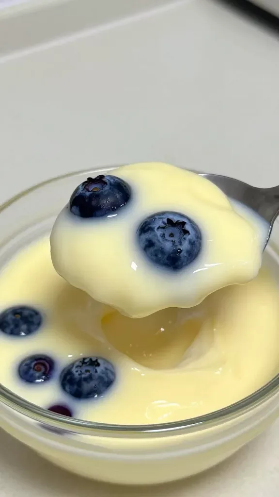 Macro side-angle of a spoonful lifted from a chilled lemon blueberry custard in a clear glass bowl: smooth, velvety custard with a uniform light-lemon hue, studded with softened whole blueberries releasing rich indigo juices; the spoon reveals creamy thickness without curdling; background softly blurred kitchen counter, neutral tones, natural soft light, no garnishes, no crust, no extra elements, no text.